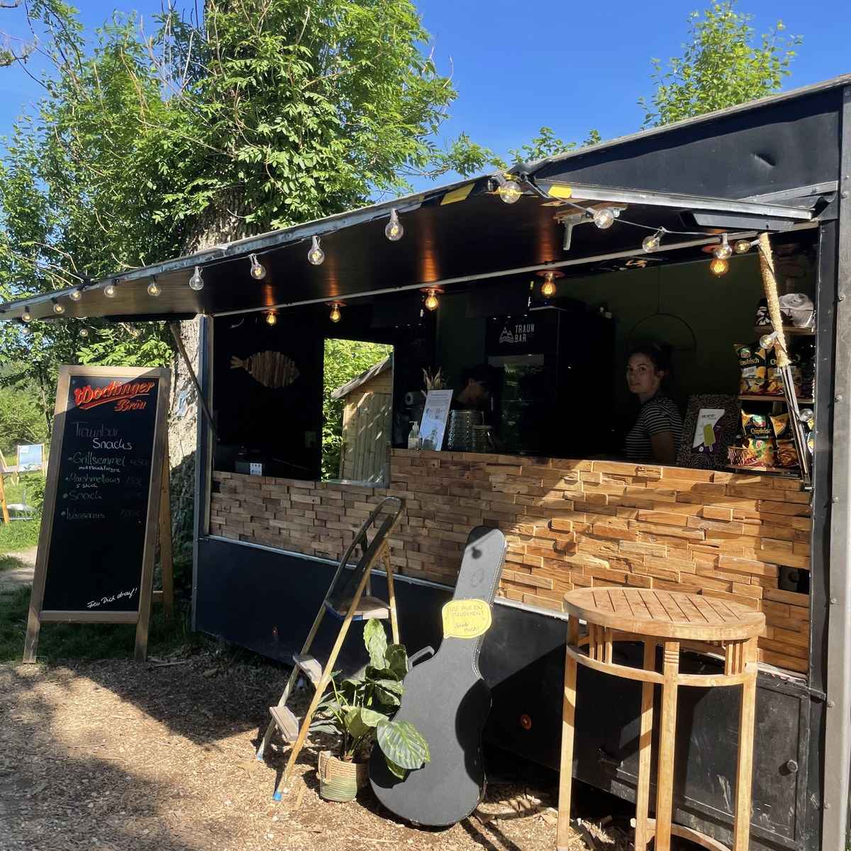Die TRAUNBAR in der Sonne: Lichterkette, Holztheke und Tafel mit Snacks unter blauem Himmel