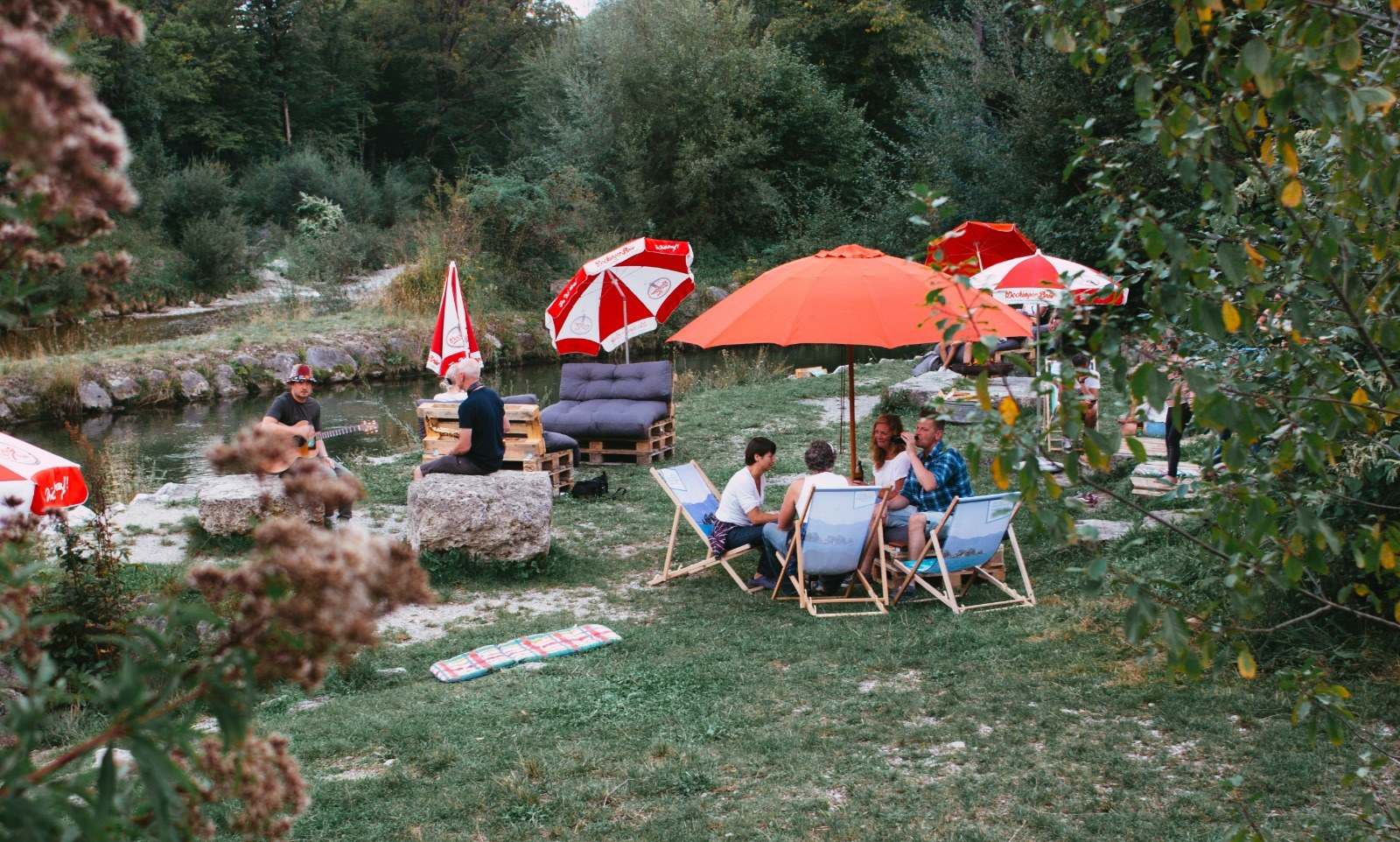 Paletten-Lounge und orange Sonnenschirme auf der Wiese am Fluss