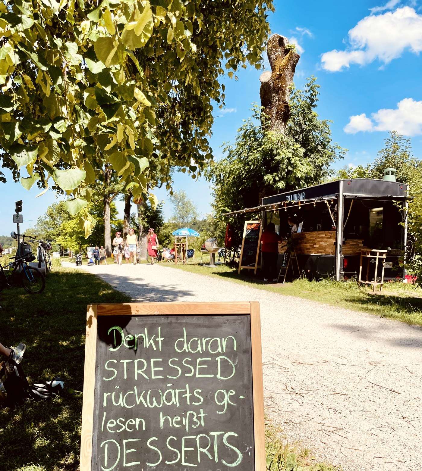 Der Kiesweg zur TRAUNBAR mit Kreidetafel „Stressed rückwärts gelesen heißt Desserts“ und der TRAUNBAR im Hintergrund